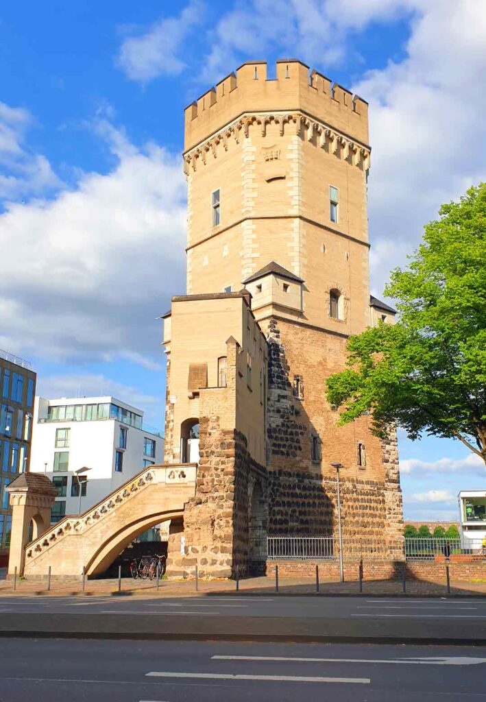 Der mittelalterliche Bayenturm aus hellem Stein mit einer Außentreppe an einer Straße in Köln unter blauem Himmel mit weißen Wolken.