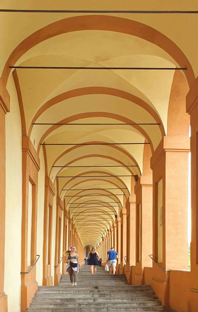 Blick die Stufen des Portico di San Luca in Bologna hinauf, einem UNESCO-Weltkulturerbe, mit den charakteristischen gelben und terracottafarbenen Rundbögen und wenigen Spaziergängern.