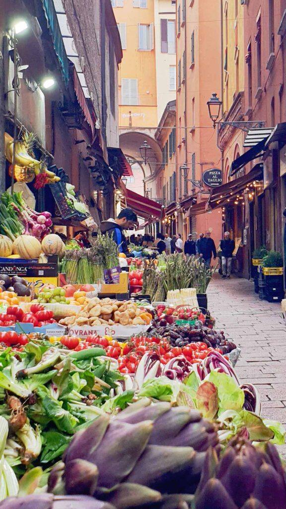 Marktstand mit frischem Gemüse und Obst, darunter Artischocken und Spargel, in der engen Gasse Via Pescherie Vecchie in Bologna, mit Passanten und terracottafarbenen Gebäuden im Hintergrund.