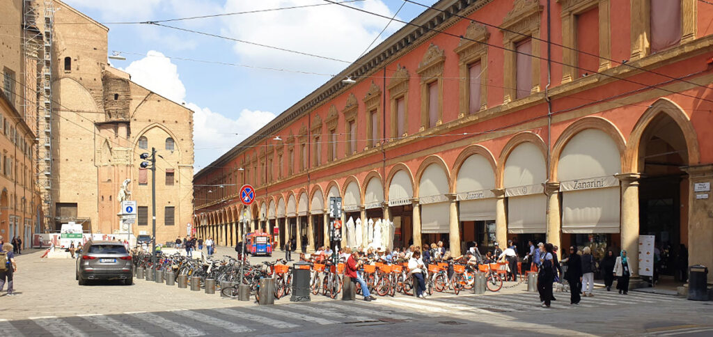 Rote Fassade des historischen Palazzo dell'Archiginnasio mit überdachten Arkaden und geparkten Fahrrädern an der Piazza Galvani hinter der Basilika San Petronio in Bologna.