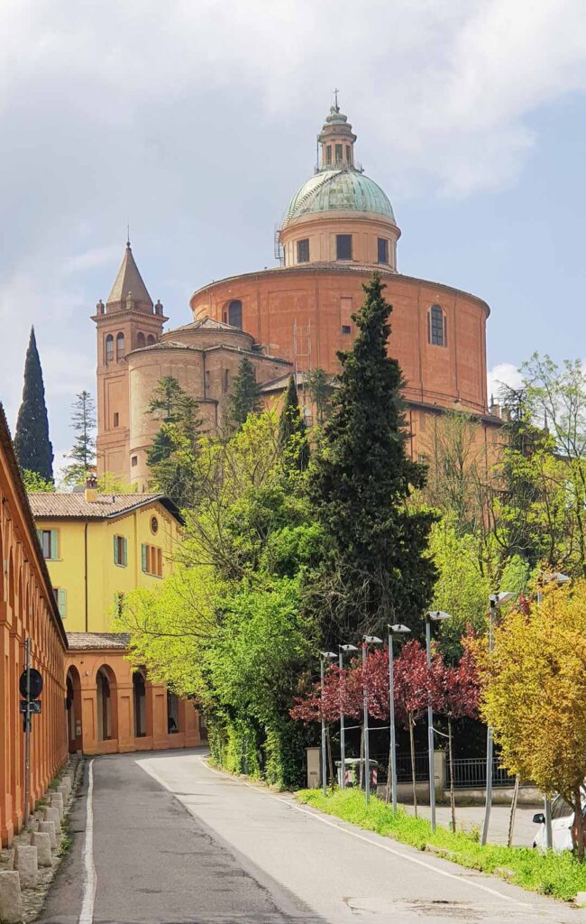 Blick von einer Straße mit den überdachten Arkaden auf die terracottafarbene Kuppel des Santuario della Madonna di San Luca in Bologna.