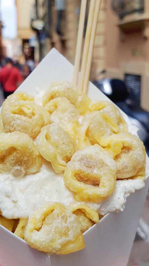 Eine Papiertüte gefüllt mit knusprig frittierten Tortellini auf einer hellen Parmesancreme mit Holzspießen.