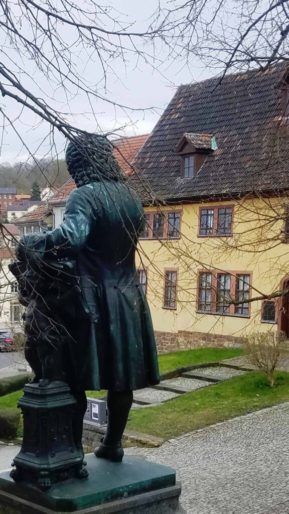Bronzene Statue des Bach-Denkmals von hinten gesehen, dahinter das gelbe historische Bachhaus in Eisenach.