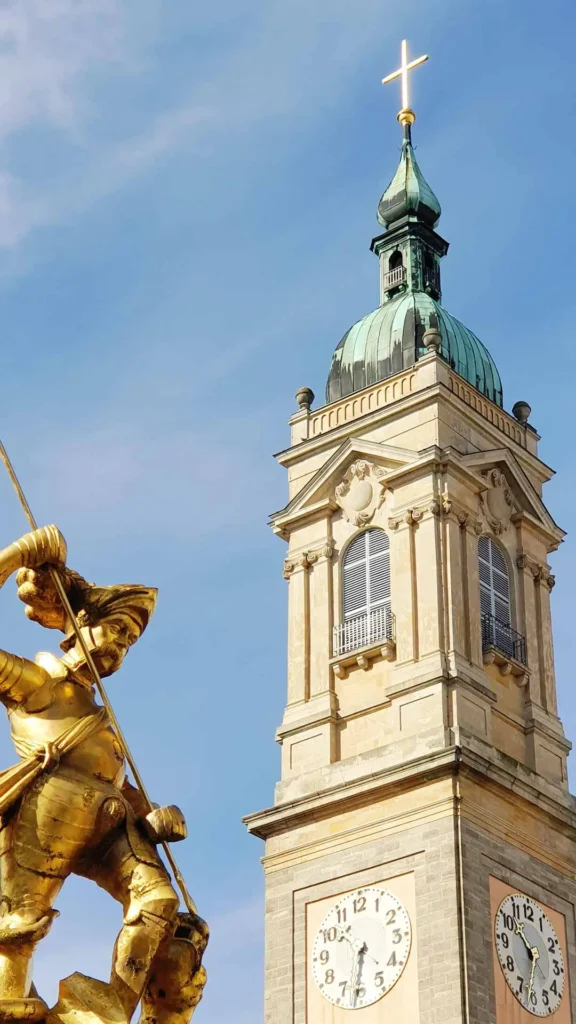 Die vergoldete Statue des Heiligen Georg auf dem Marktbrunnen in Eisenach vor dem imposanten Kirchturm der Georgenkirche unter blauem Himmel.