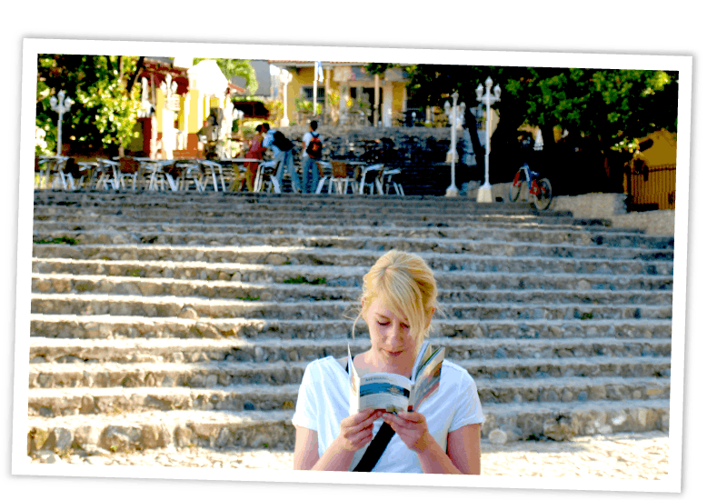 Eine junge Frau mit blonden Haaren vor einer breiten Steintreppe mit einem Café im Freien und konzentriert sich auf das Lesen eines Reiseführers.