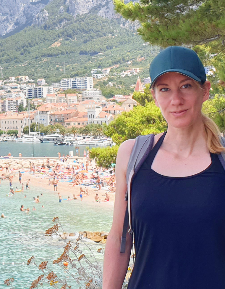 Reisebloggerin mit dunkler Cap und Rucksack im Vordergrund, im Hintergrund eine sonnige mediterrane Küstenstadt mit Sandstrand, türkisblauem Meer, Booten und Bergen.