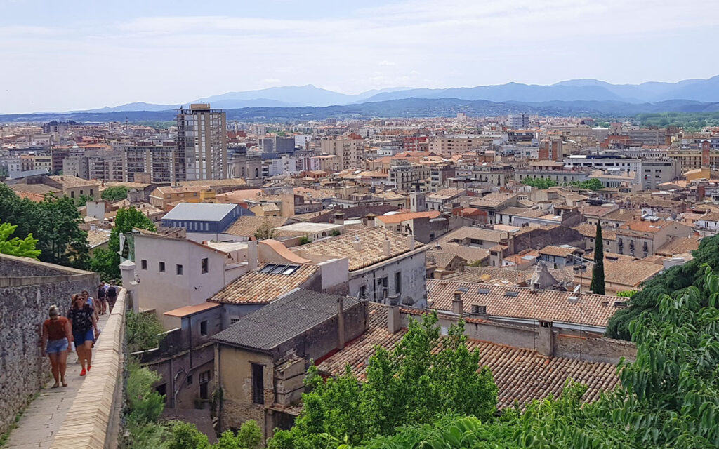 Panoramablick von der historischen Stadtmauer in Girona über die roten Terrakotta-Dächer der Altstadt bis zur modernen Skyline und den Bergen im Hintergrund.
