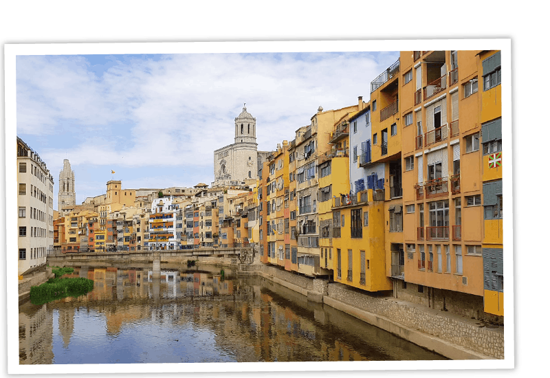 Historische bunte Häuser am Onyar-Fluss in Girona mit der Kathedrale im Hintergrund, präsentiert in einem weißen Polaroid-Bildrahmen.