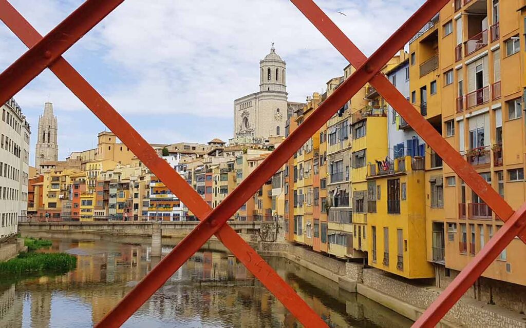 Blick auf die bunten Häuser am Fluss Onyar und die Kathedrale in Girona durch das rote Eisengitter der Eiffel-Brücke
