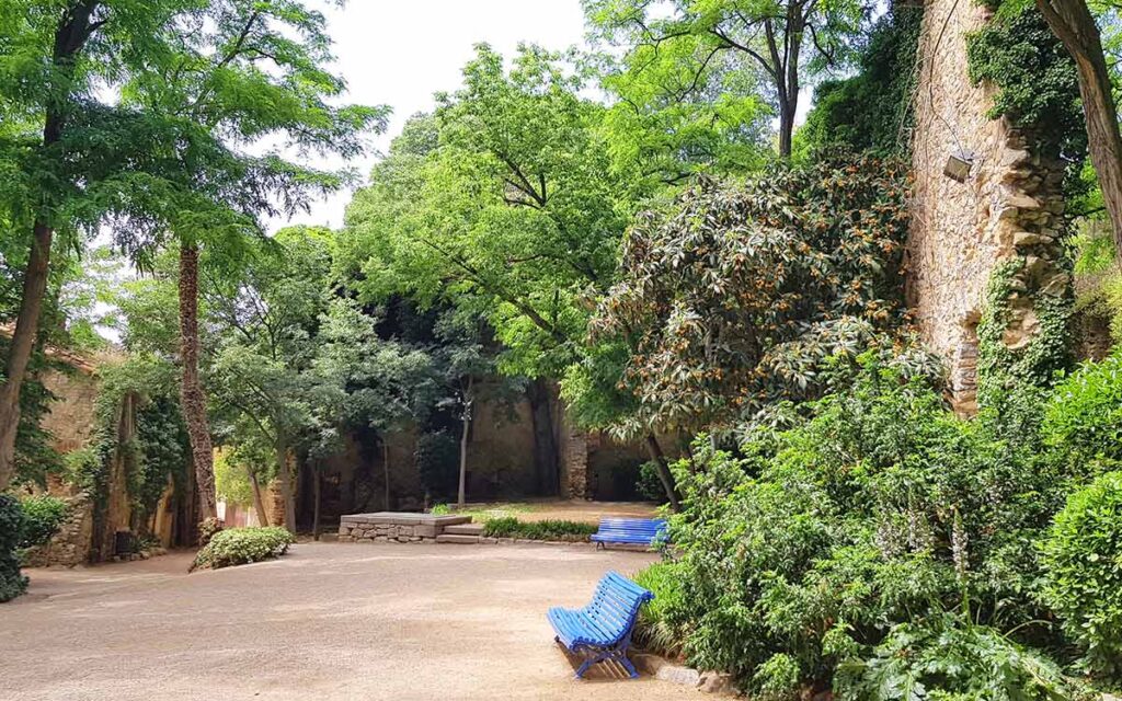Grüner, versteckter Innenhof in Girona mit alten, mit Efeu bewachsenen Steinmauern, üppigen Bäumen und zwei leuchtend blauen Parkbänken im Schatten.