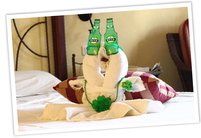 Eine kreative Handtuch-Faltung in Form eines Tieres auf einem Hotelbett, geschmückt mit einer grünen Bierflaschen-Brille und einer grünen Kleeblatt-Kette.