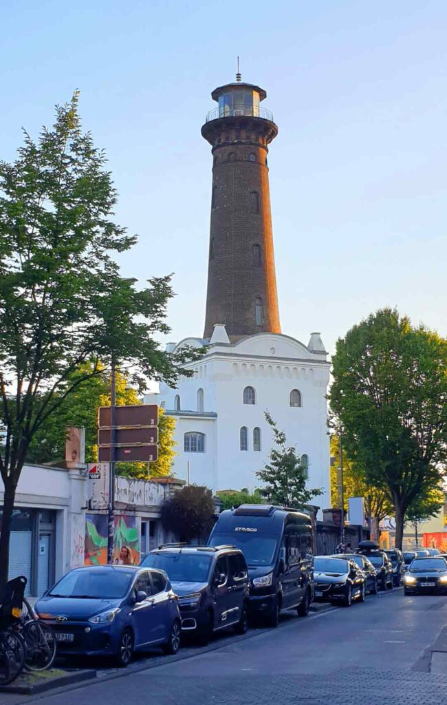 Der historische Helios-Leuchtturm aus Backstein in Köln-Ehrenfeld über einer Straße mit parkenden Autos.