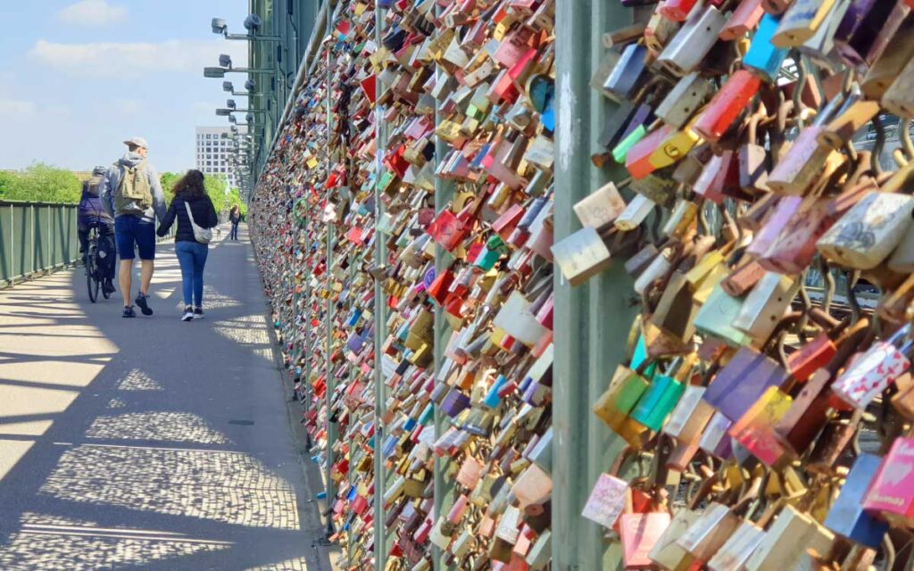 Tausende bunte Liebesschlösser am Gitter der Hohenzollernbrücke in Köln mit Fußgängern auf dem Gehweg.
