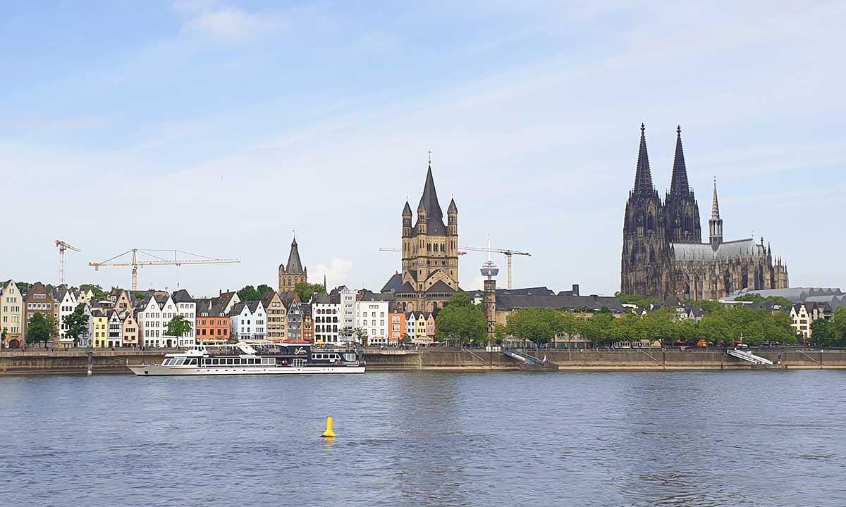 Panorama-Blick über den Rhein auf die bunten Giebelhäuser der Kölner Altstadt, die Kirche Groß St. Martin und den Kölner Dom unter leicht bewölktem Himmel.