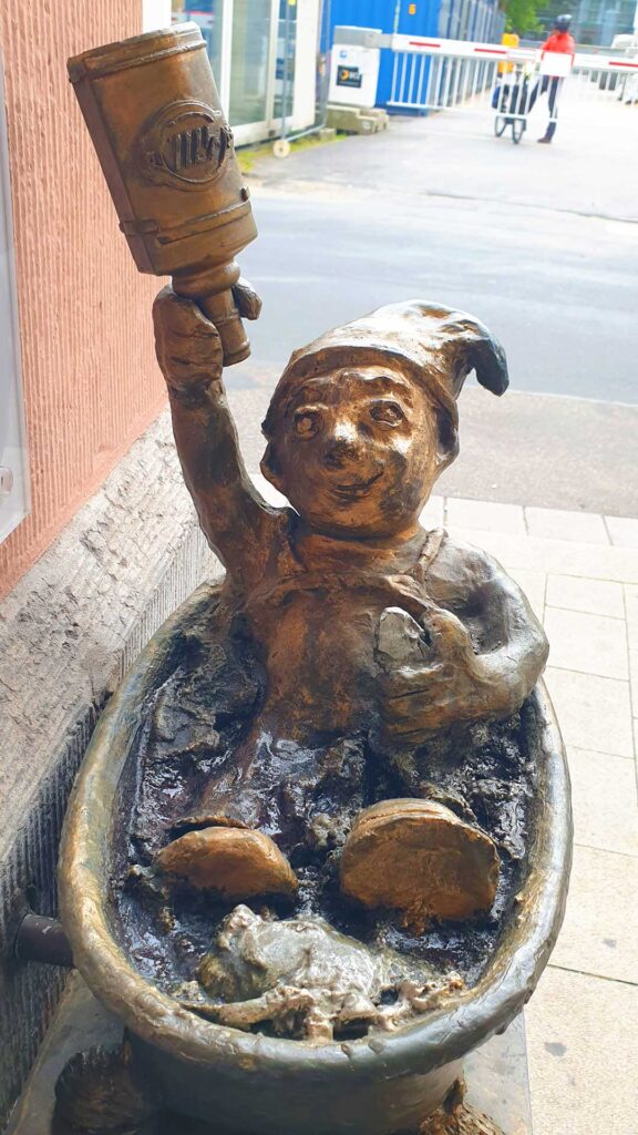 Eine kleine Bronzefigur eines Kölner Heinzelmännchens in einer Waschwanne, das eine Flasche 4711 Echt Kölnisch Wasser hochhält.