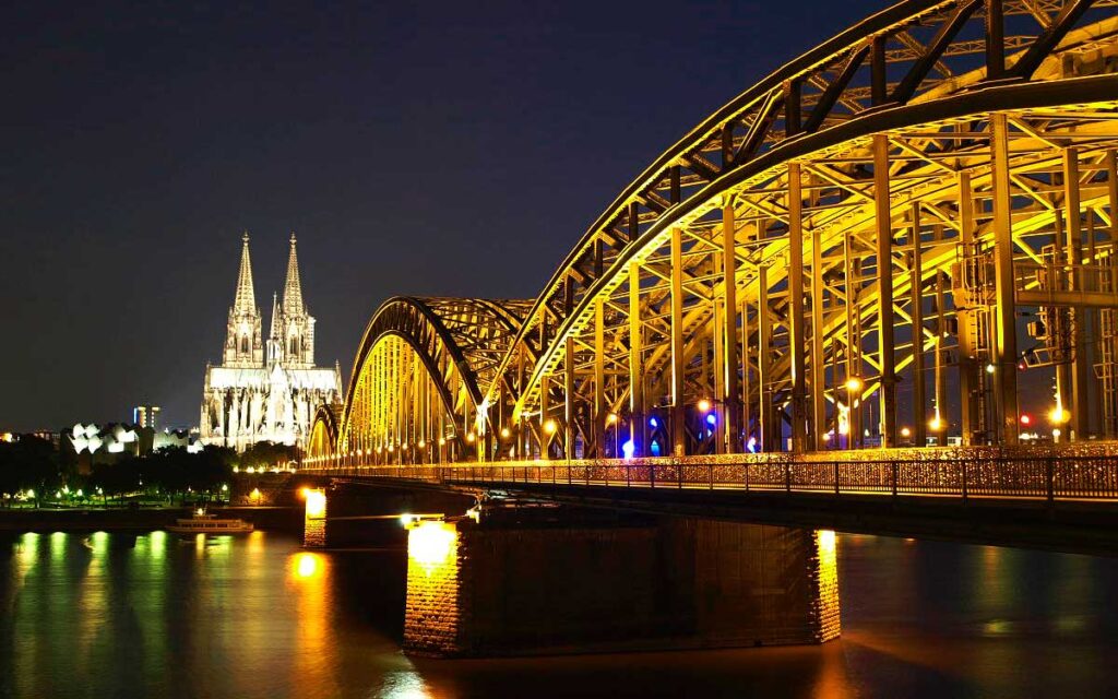 Der beleuchtete Kölner Dom und die golden strahlende Hohenzollernbrücke bei Nacht mit Spiegelungen auf dem Rhein.