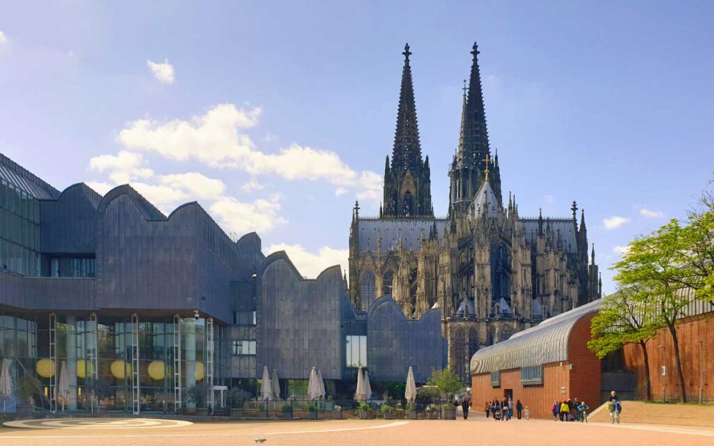 Blick über den Heinrich-Böll-Platz auf den Kölner Dom hinter der modernen Architektur des Museums Ludwig bei blauem Himmel.