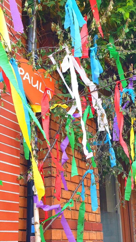 Detailaufnahme einer Maibaum-Birke in Köln mit einem roten Herz mit der Aufschrift Paula und bunten Bändern vor einer Backsteinwand.