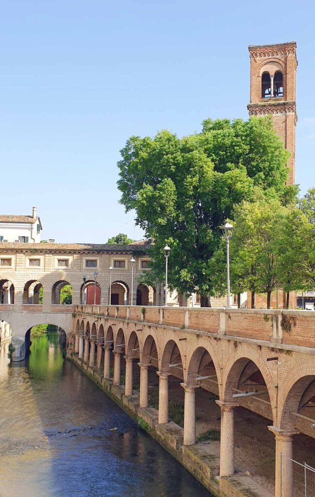 Blick über den Rio-Kanal in Mantua auf die doppelstöckigen Pescherie-Loggien von Giulio Romano und den Torre dell'Orologio unter blauem Himmel.