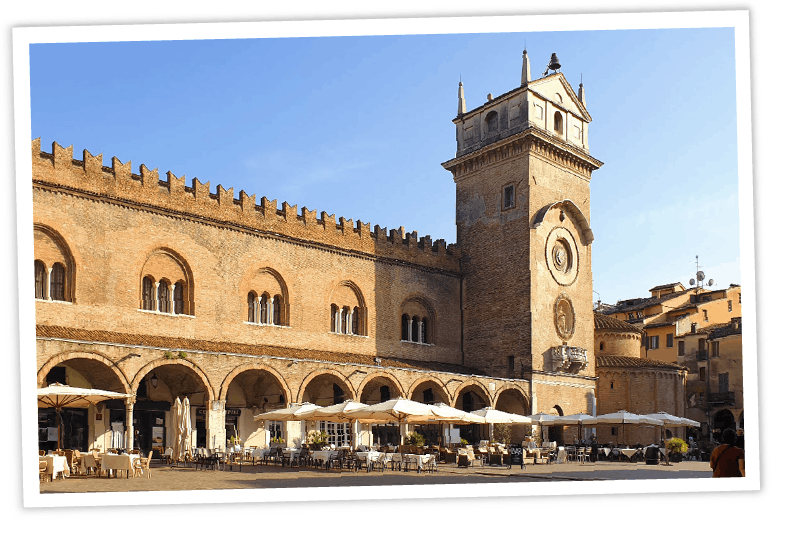 Sonniger Blick auf die Piazza delle Erbe in Mantua (Italien) mit dem historischen Palazzo della Ragione, dem Uhrturm Torre dell'Orologio und einem Straßencafé unter Sonnenschirmen.