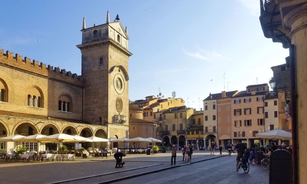 Ein Panoramablick auf den historischen Piazza delle Erbe in Mantua, Italien, mit dem mittelalterlichen Palazzo della Ragione (links) und dem Torre dell'Orologio (Uhrturm, Mitte) unter einem klaren blauen Himmel.