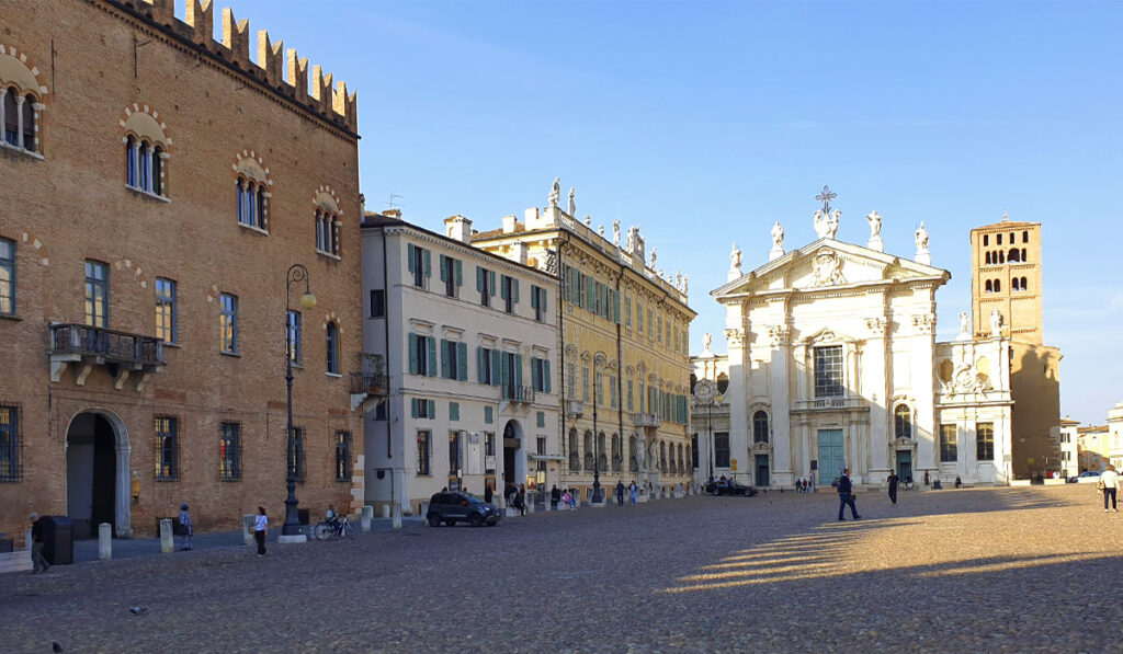 Blick über den gepflasterten Piazza Sordello in Mantua auf die helle Marmorfassade des Doms von Mantua (Cattedrale di San Pietro) und den romanischen Glockenturm unter blauem Himmel.