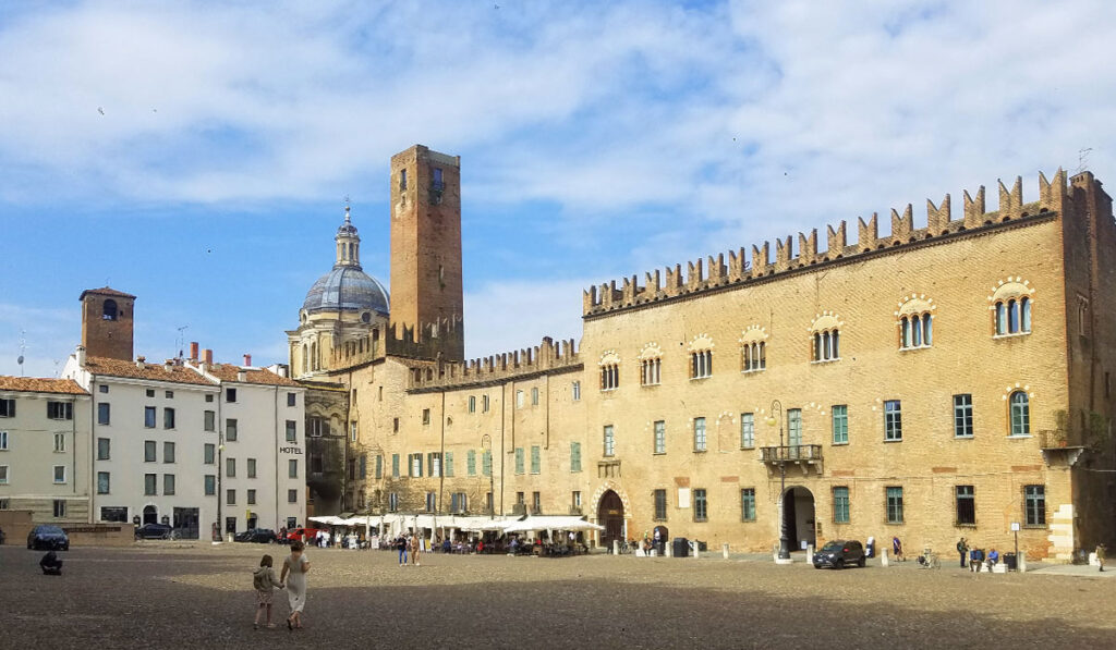 Der weitläufige, gepflasterte Piazza Sordello in Mantua mit der imposanten Backsteinfassade des Palazzo Ducale, dem Torre della Gabbia und der Kuppel der Basilica di Sant'Andrea im Hintergrund.
