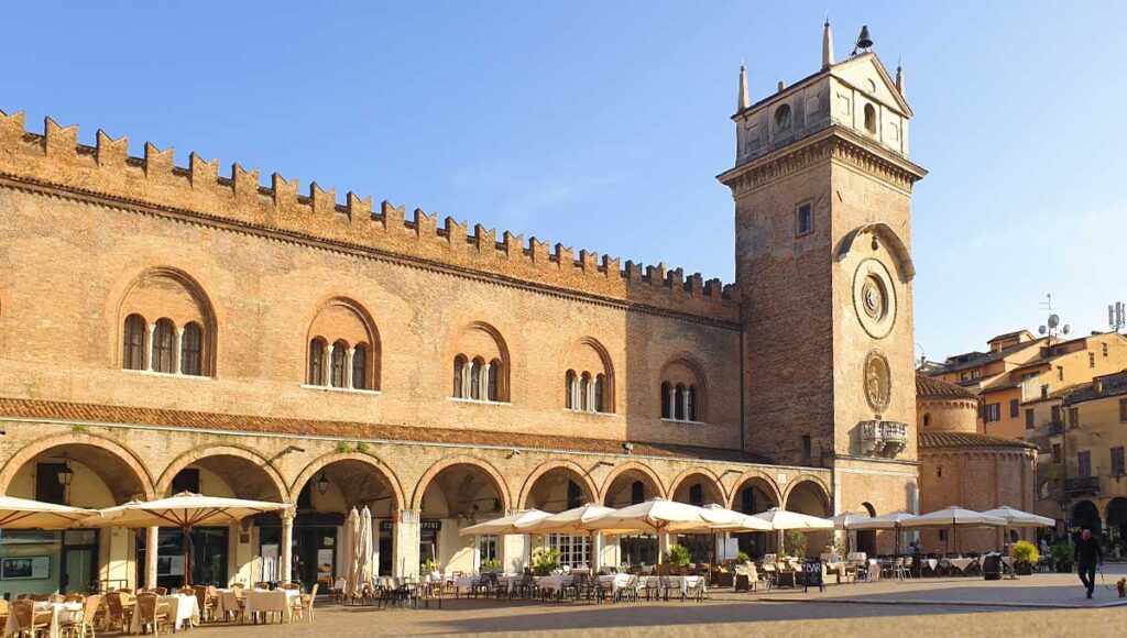 Historische Gebäude und der Uhrturm an der Piazza delle Erbe in Mantua mit gemütlichen Cafés unter Sonnenschirmen.
