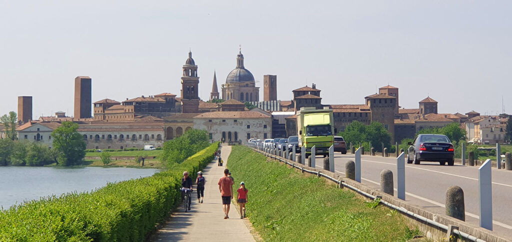 Panorama der historischen Skyline von Mantua, Italien, mit dem Castello di San Giorgio und Kirchtürmen, gesehen über einen See mit einem Radweg im Vordergrund.