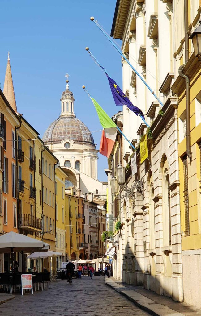 Blick durch die schmale, gepflasterte Via Calvi in Mantua mit farbenfrohen Gebäuden auf die große barocke Kuppel der Basilica di Sant'Andrea unter blauem Himmel.