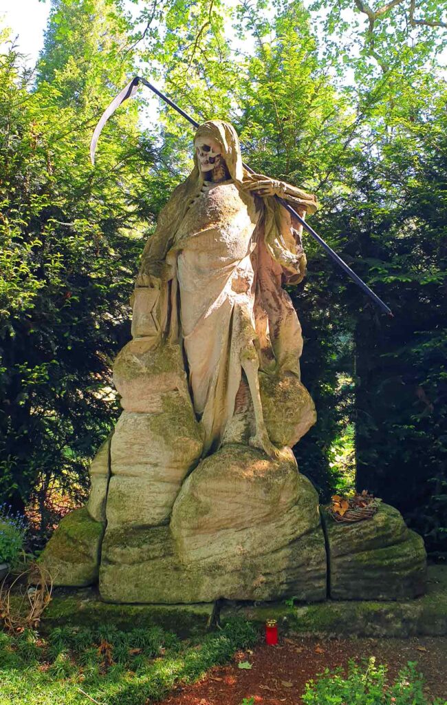 Die historische Steinstatue des Sensenmanns mit einer großen Sense vor dichten grünen Hecken auf dem Melatenfriedhof in Köln.
