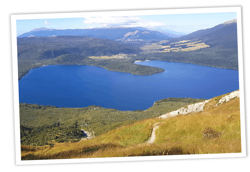 Weitwinkelaufnahme vom Mount Robert-Wanderweg auf den strahlend blauen Lake Rotoiti, umgeben von dichten Bergen, mit einem Tussock-Hang und schmalem Pfad im Vordergrund.