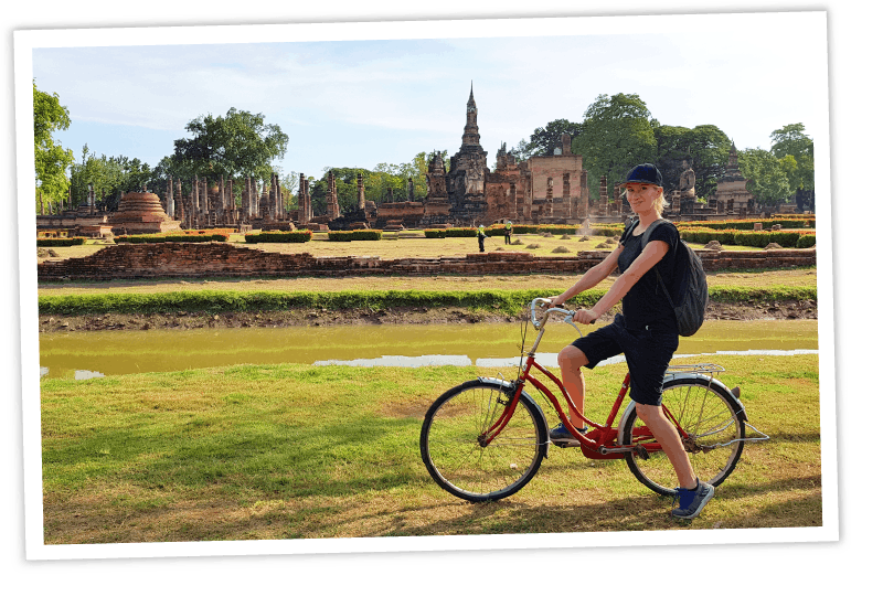 Reisebloggerin mit Rucksack auf einem roten Retro-Fahrrad vor der historischen Kulisse der alten Tempelruinen im Sukhothai Historical Park in Thailand, umgeben von Grünflächen und einem Wassergraben.