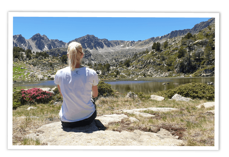 Reisebloggerin von hinten auf einem Felsen sitzend, den Blick auf einen Bergsee und ein Bergpanorama in einer sonnigen alpinen Landschaft gerichtet.