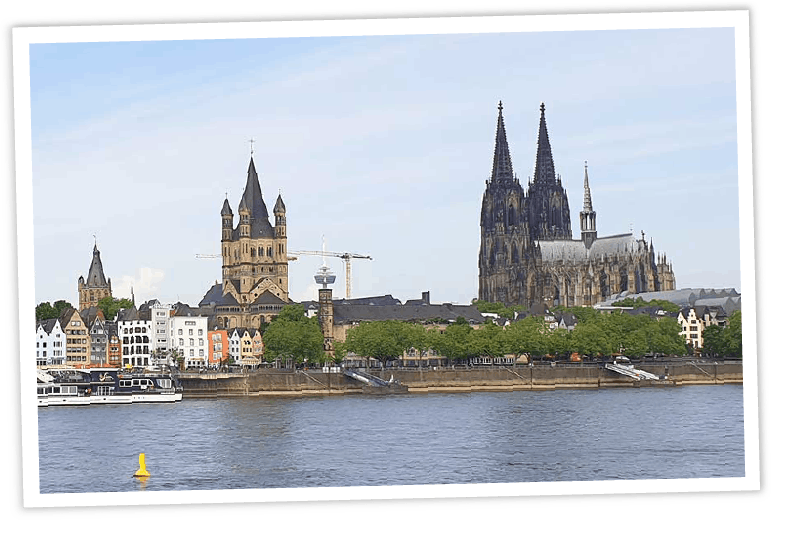 Teaserbild für Deutschland Reisetipps: Ein Panorama-Blick über den Rhein auf die Kölner Altstadt mit Groß St. Martin und dem Dom, gestaltet im Stil eines schräg liegenden Fotos mit weißem Rand.