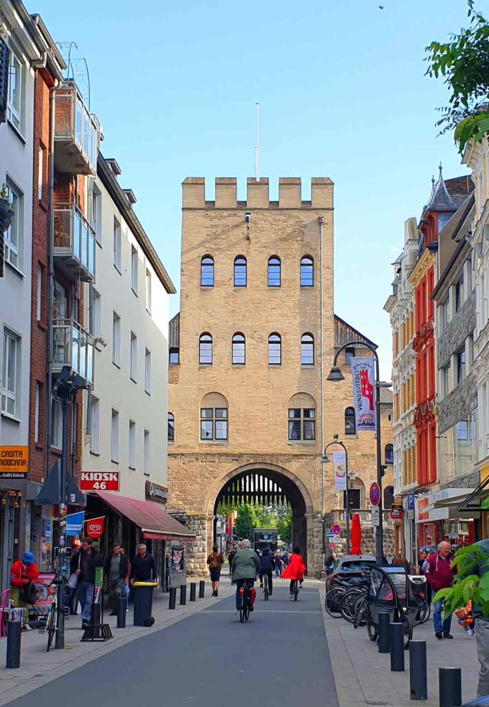 Blick die belebte Severinstraße hinunter auf das historische Stadttor Severinstorburg im Kölner Severinsviertel bei blauem Himmel.
