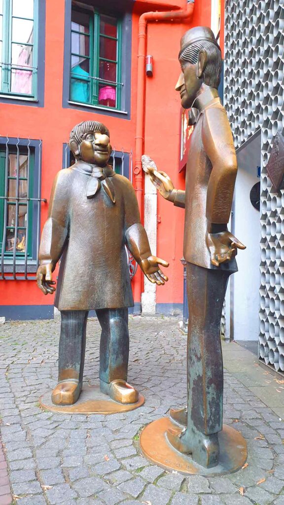 Die Bronzestatuen von Tünnes und Schäl auf dem Kopfsteinpflaster vor einer roten Hausfassade in der Kölner Altstadt.