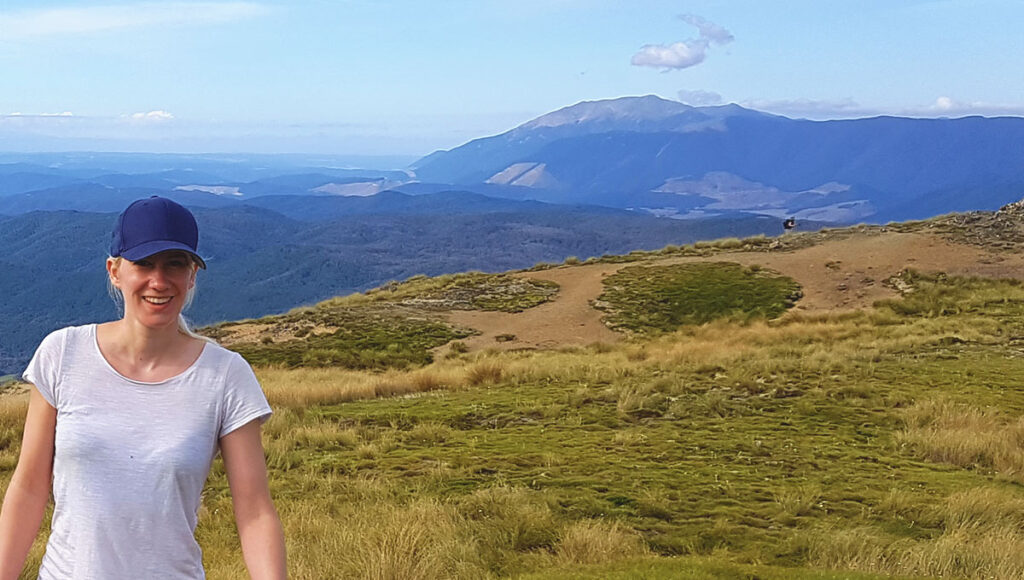 Eine Wanderin mit blauer Mütze und weißem Shirt steht lächelnd auf dem offenen Plateau des Mount Robert mit Blick auf die Berglandschaft.